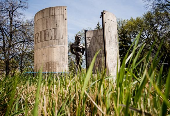 Statue de Louis Riel placée entre deux grandes structures de béton courbées gravées du nom "RIEL", entourée d’arbres et vue à travers l’herbe haute au premier plan, sous un ciel bleu.