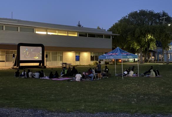 Assises sur des couvertures et des chaises, des personnes sont rassemblées sur la pelouse du campus de l'université pour une soirée cinéma en plein air, un film projeté sur un écran gonflable à la tombée du jour.