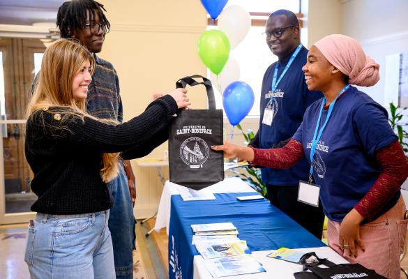 Deux membres de l’équipe ambassadrice accueillent deux visiteurs à une table d’information, leur remettant un sac promotionnel, tandis que des brochures et des ballons sont visibles.