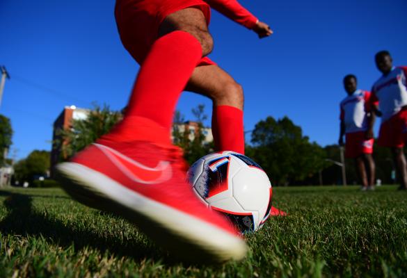 Un joueur de soccer dribblant un ballon sur un terrain de gazon, avec d’autres joueurs en arrière-plan sous un ciel bleu dégagé.