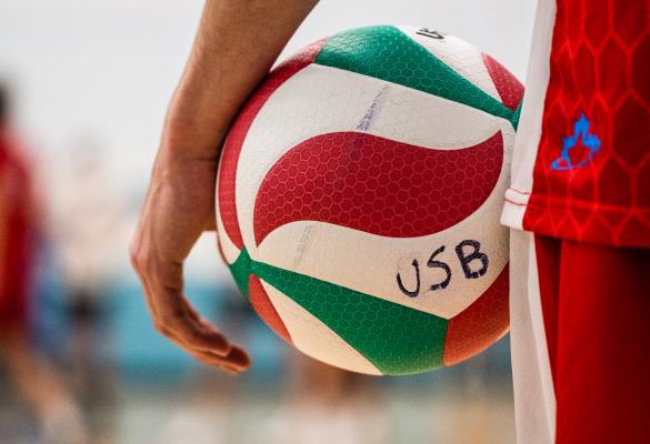 Un joueur de volleyball en uniforme rouge tenant un ballon en gros plan. Le terrain de gymnase est flou en arrière plan..