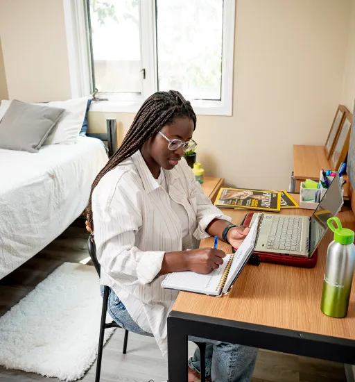 Une étudiante écrit dans son cahier à son bureau, avec un ordinateur portable, des revues, un lit et une fenêtre en arrière-plan.