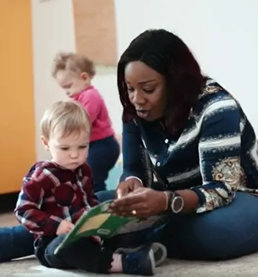 Une femme assise par terre regarde un livre avec un tout-petit à sa droite. Un autre enfant en bas âge est debout derrière eux. 