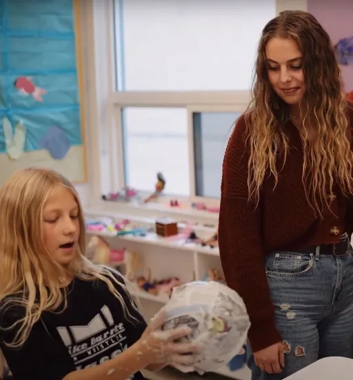 Une femme debout entre deux jeunes filles assises à une table regarde à sa droite celle qui tient un objet en papier mâché. 