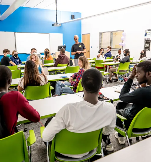 Une vingtaine de personnes assises à des tables dans une salle de classe interagissent entre elles et avec un homme qui se tient debout parmi elles.