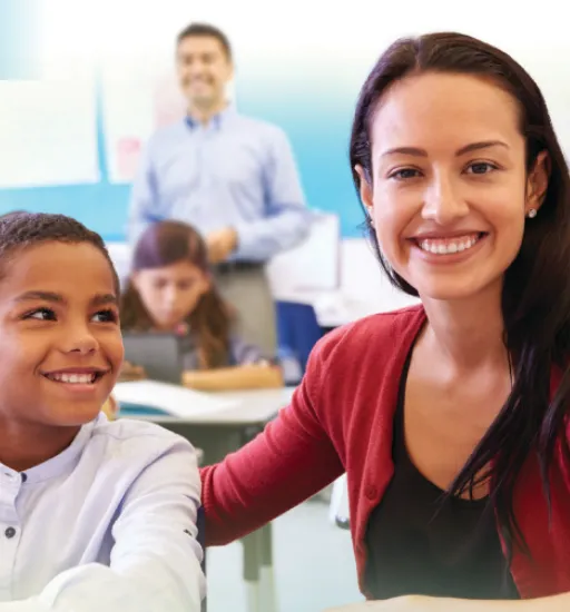 Un enfant assis dans une salle de classe tient une tablette et sourit à une jeune femme assise à ses côtés, qui sourit en regardant l’objectif.  