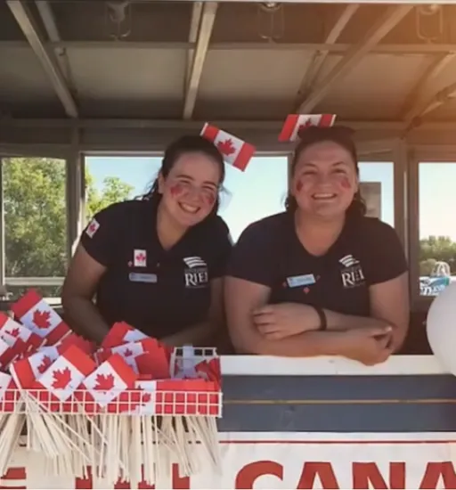 Deux personnes souriant s’appuient sur le comptoir d’un kiosque, derrière un présentoir de petits drapeaux canadiens.