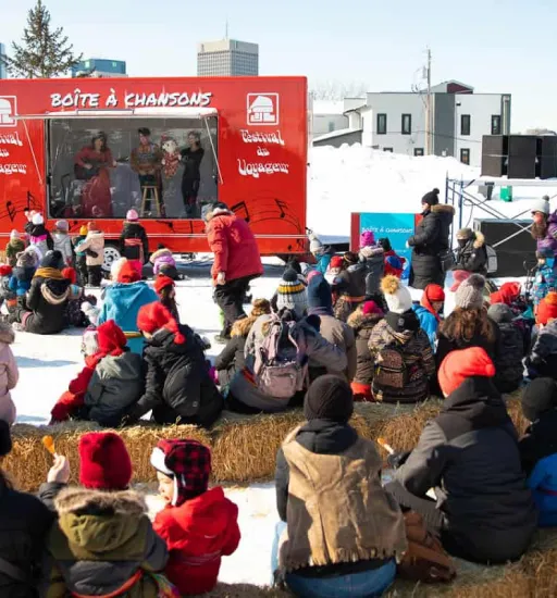 Une trentaine de personnes assises sur des ballots de foin dans la neige regardent un spectacle de musique dehors en hiver.