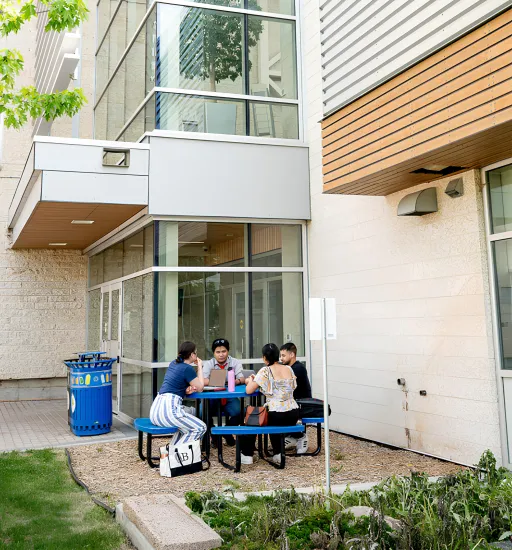 Quatre personnes sont assises à une table de piquenique près d’une porte d’entrée du campus et discutent.