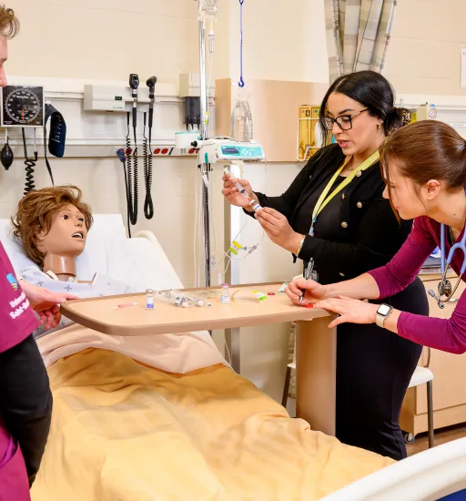 Une professeure démontre une technique de seringue à quelques personnes étudiantes autour d’un lit d’hôpital dans lequel est couché un mannequin.