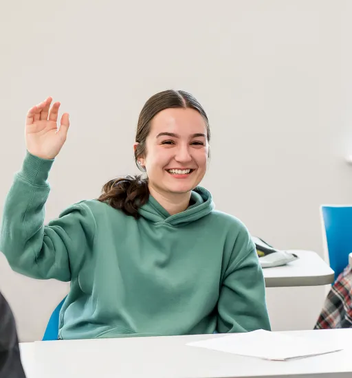 Une élève souriante, vêtue d'un chandail à capuchon turquoise, est assise à son pupitre, la main levée pour poser une question. 