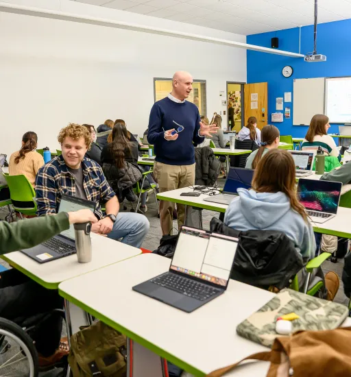 Un homme se tient debout au milieu d'une salle de classe bien remplie, ses lunettes à la main, et discute avec un groupe d'élèves.