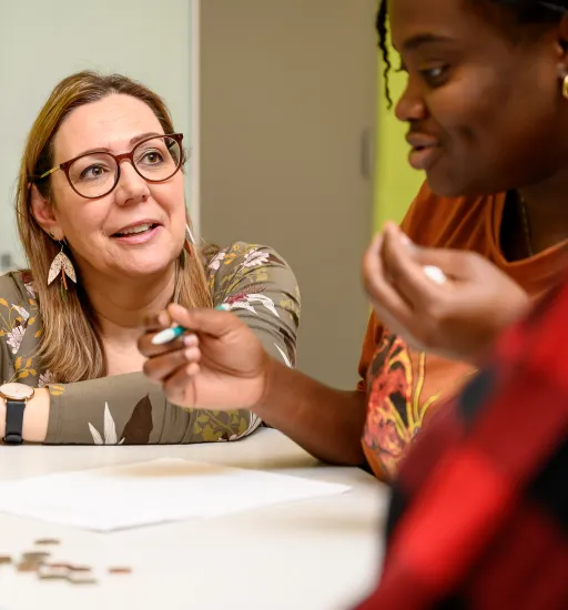 Une prof, avec des lunettes et des boucles d'oreilles en forme de fleurs, est accroupie à côté d'une étudiante du programme EJE assise à une table. Les deux discutent activement tout en regardant divers items d'apprentissage sur la table.