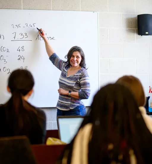 Trois personnes vues de dos sont assises dans une salle de classe et écoutent une prof à l’avant qui leur regarde en souriant.