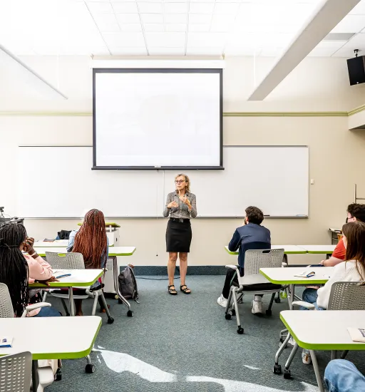 Dans une salle de classe, une femme est debout devant un tableau blanc et parle à neuf étudiantes et étudiants assis à des tables que l’on voit de dos.  