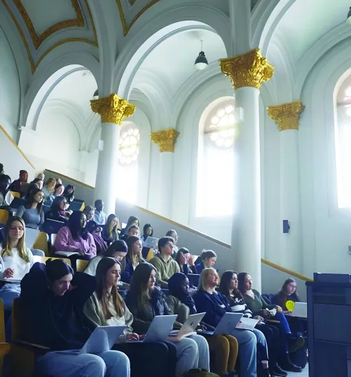 Un auditorium rempli de personnes étudiantes, plusieurs avec un ordinateur sur les genoux, regarde vers l’avant.