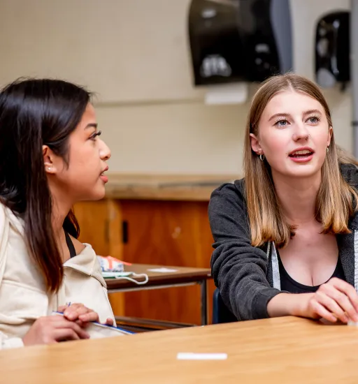 Deux jeunes femmes assises à une table discutent. Celle de gauche a les cheveux noirs, celle de droite, les cheveux châtains.  