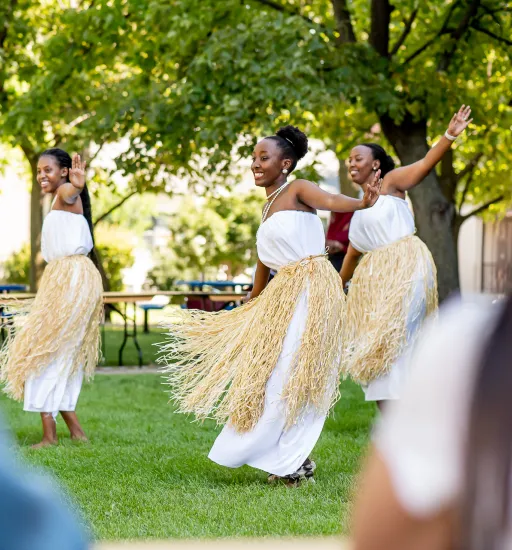 Trois femmes en robes blanches et beiges exécutent une danse traditionnelle en plein air devant des spectateurs qu'on ne voit que de dos.