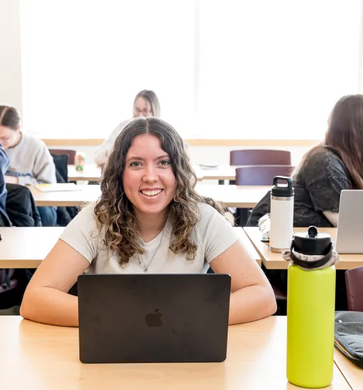 Une étudiante aux longs cheveux ondulés est assise à une table dans une salle de classe, son ordinateur portable posé devant elle. Elle sourit à la caméra.