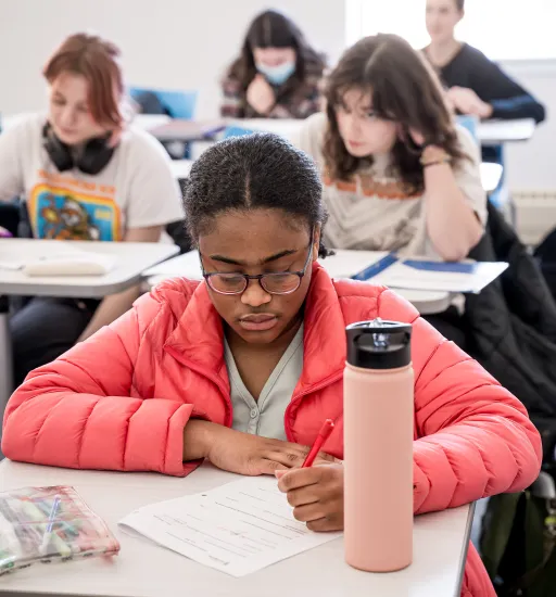 Des personnes assises à des tables. Une étudiante, à l’avant-plan, écrit. Les personnes derrière elle sont floues. 