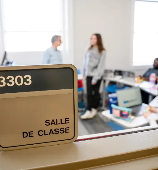 Une fenêtre donne sur une petite salle de classe, où on voit deux personnes debout qui discutent tandis que quelques autres sont assises et écoutent.