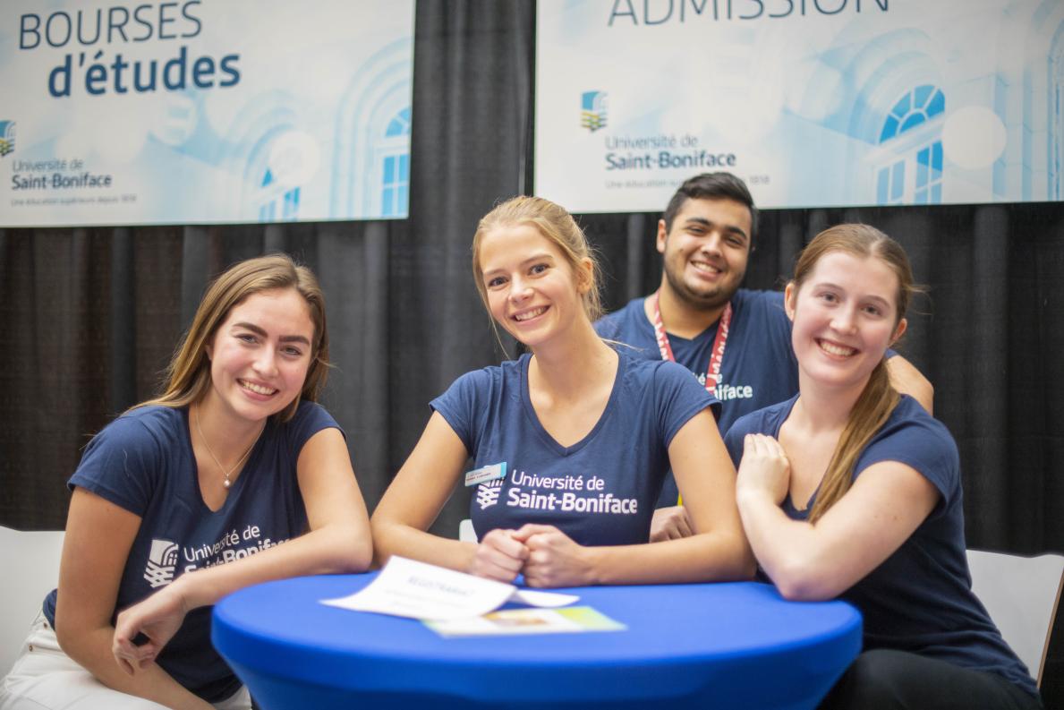 Quatre personnes assises à une table d’information de l’Université de Saint-Boniface, avec des bannières sur les bourses et l’admission en arrière-plan.