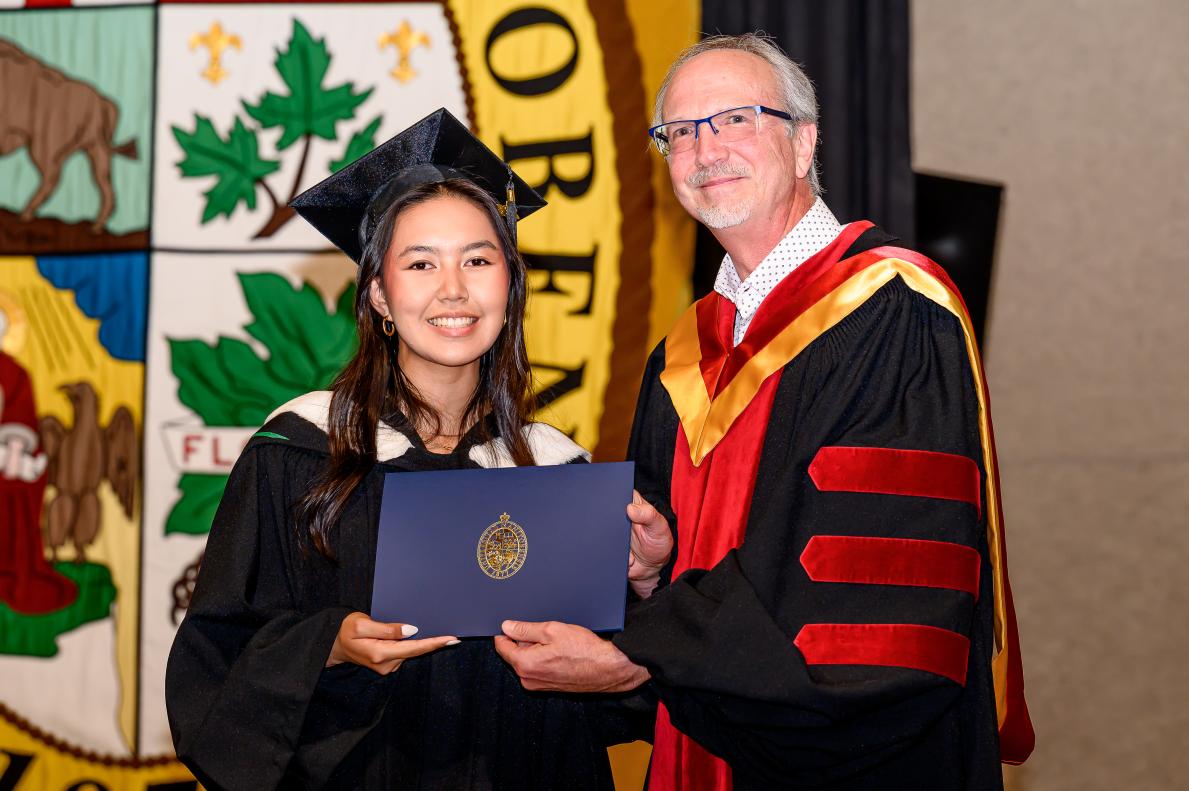 Sur une scène décorée, une personne en toge et mortier tient fièrement un diplôme, et à son côté une personne en robe universitaire noire, rouge et or.