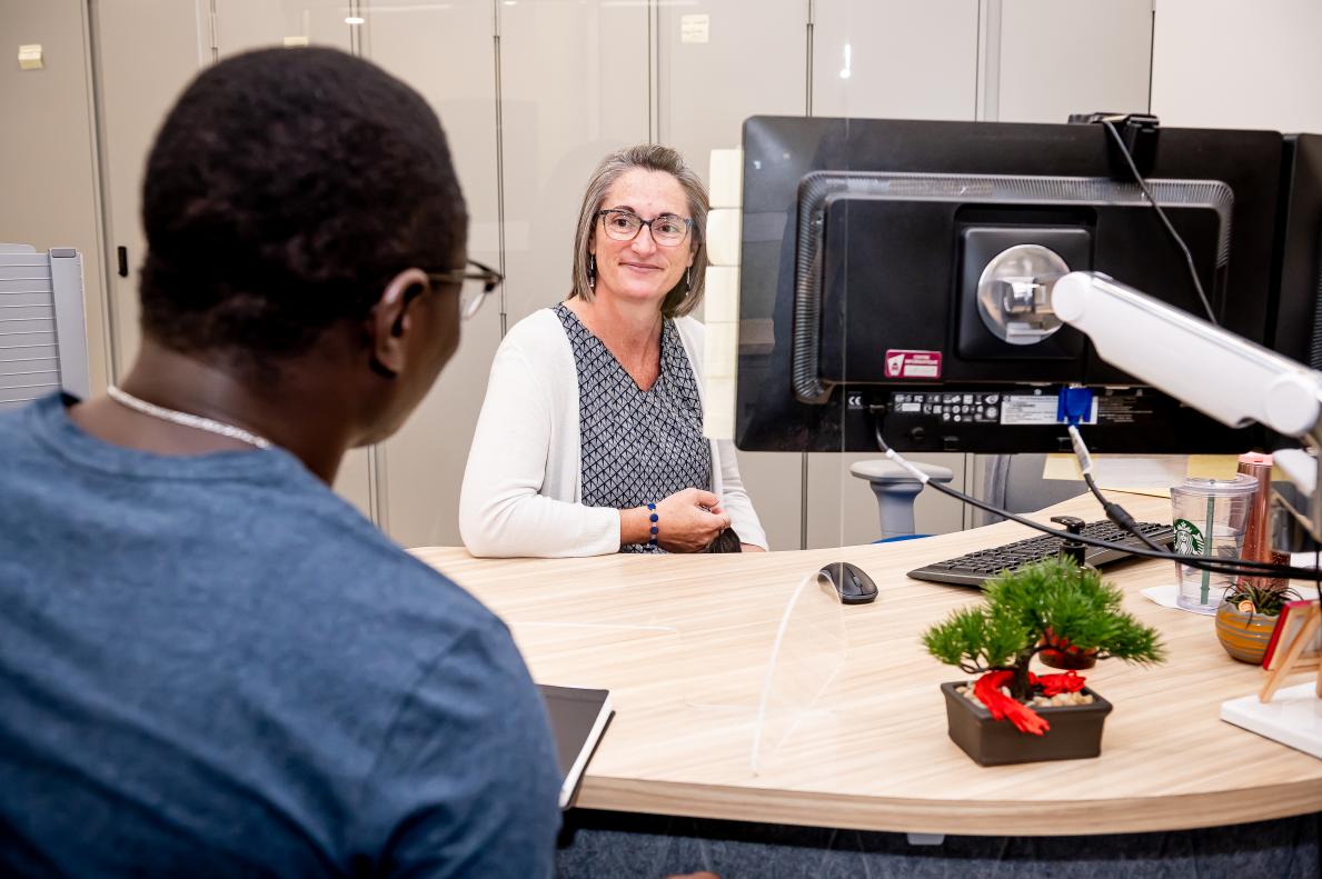 Une dame souriante accueille à son poste de travail un étudiant.