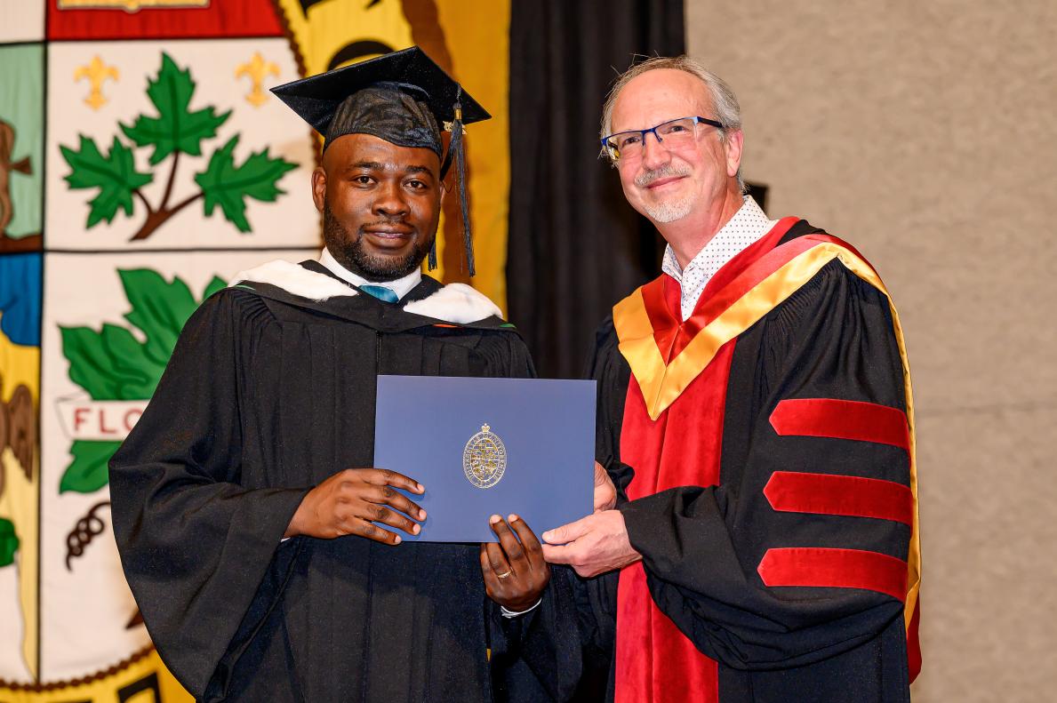 Sur une scène décorée, une personne en toge et mortier tient fièrement un diplôme, et à son côté une personne en robe universitaire noire, rouge et or.