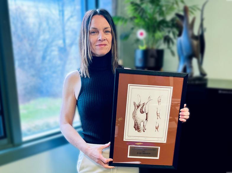 Debout près d'une fenêtre, Julie Arnaud sourit fièrement tenant un cadre contenant une illustration artistique du prix Ronald-Duhamel.