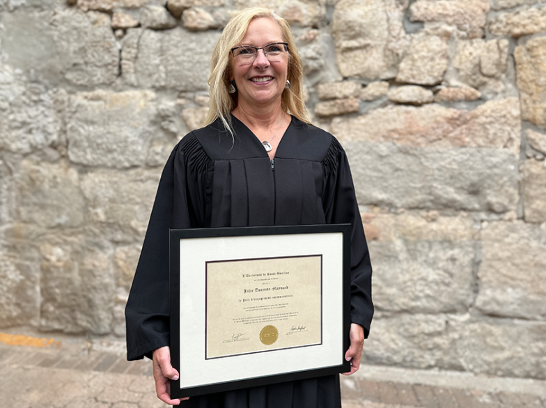 Julie Turenne-Maynard sourit en toge devant un mur de pierre, tenant le Prix de l’engagement communautaire remis par l’Université de Saint-Boniface.