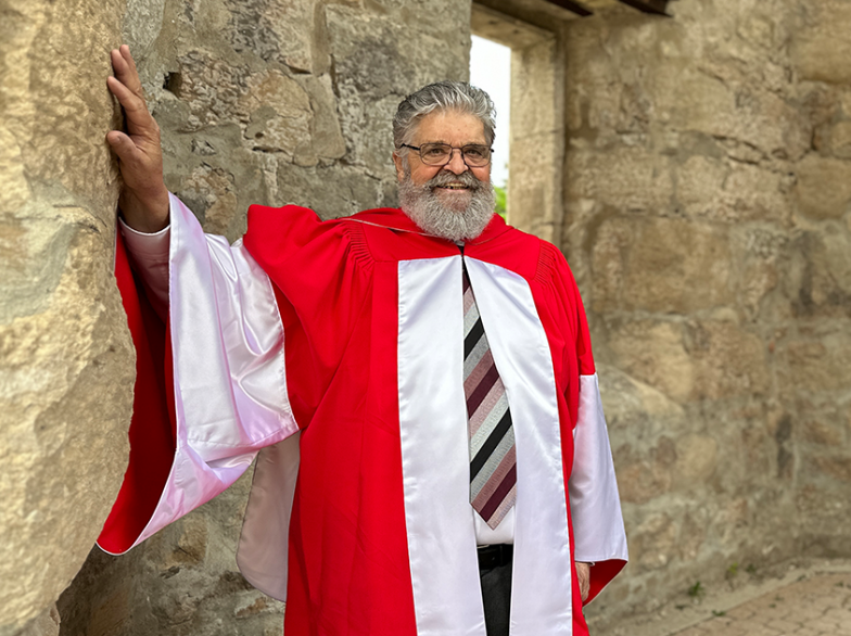 Léo Robert sourit en toge rouge de docteur honoris causa devant un mur de pierre, après avoir reçu une distinction de l’Université de Saint-Boniface.