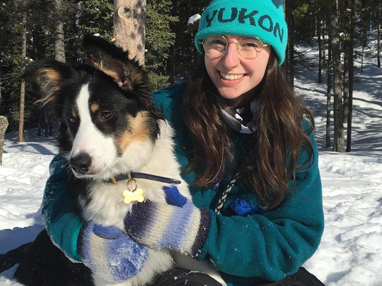 Marguerite Tölgyesi souriante en plein air, assise dans la neige avec un chien dans ses bras, portant une tuque turquoise « Yukon ».