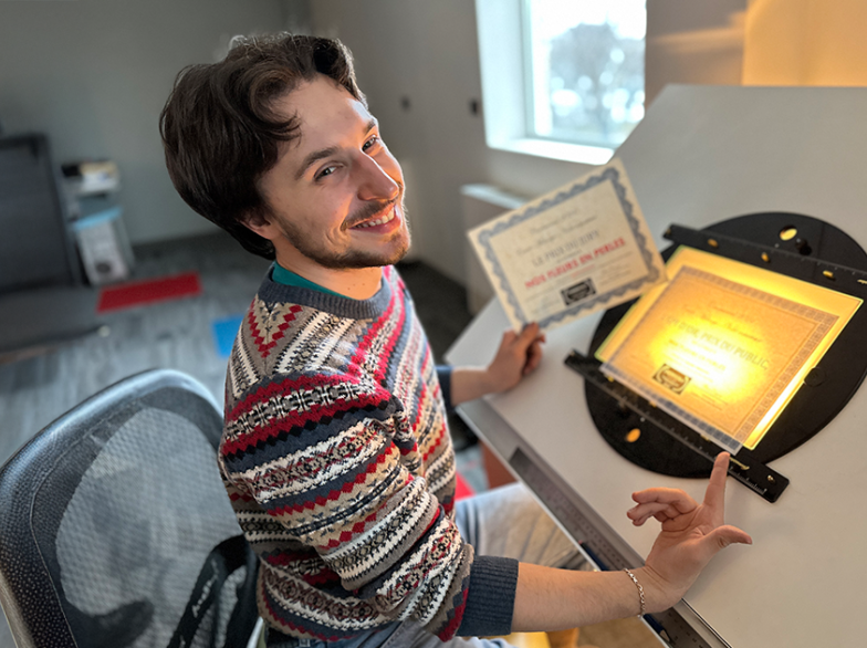 Maxime Kornachuk sourit devant sa table lumineuse, tenant fièrement un certificat pour son court métrage primé Nos fleurs en perles.