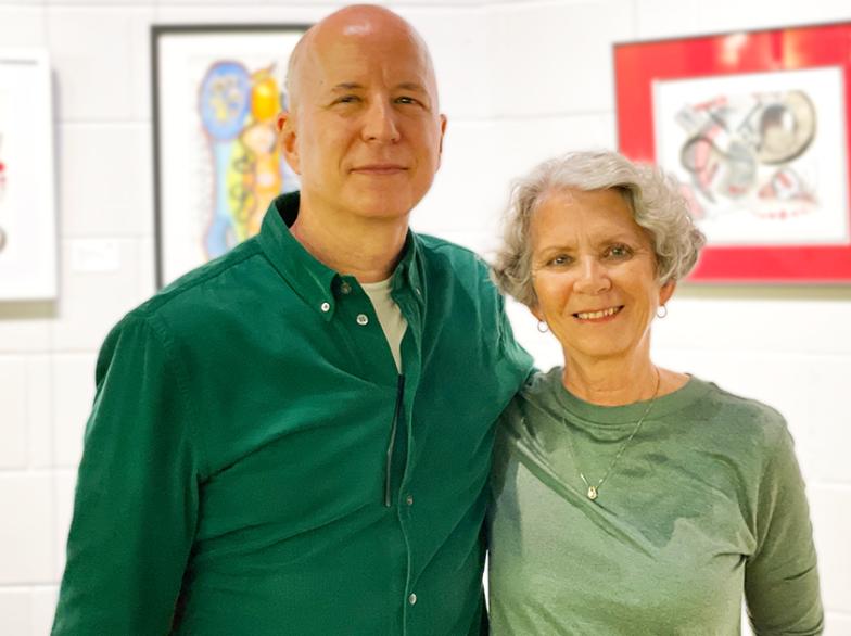 Paulette et Jacques Desaulniers posent côte à côte dans une galerie, entourés de leurs œuvres colorées exposées à l’Université de Saint-Boniface.