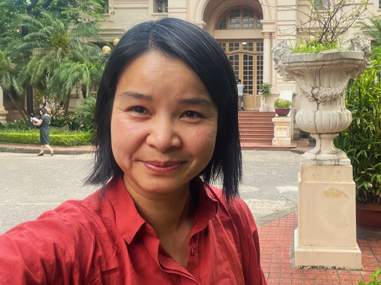 La professeure Phi-Vân Nguyen sourit devant un bâtiment historique au Vietnam, entourée de végétation tropicale et de fleurs colorées.