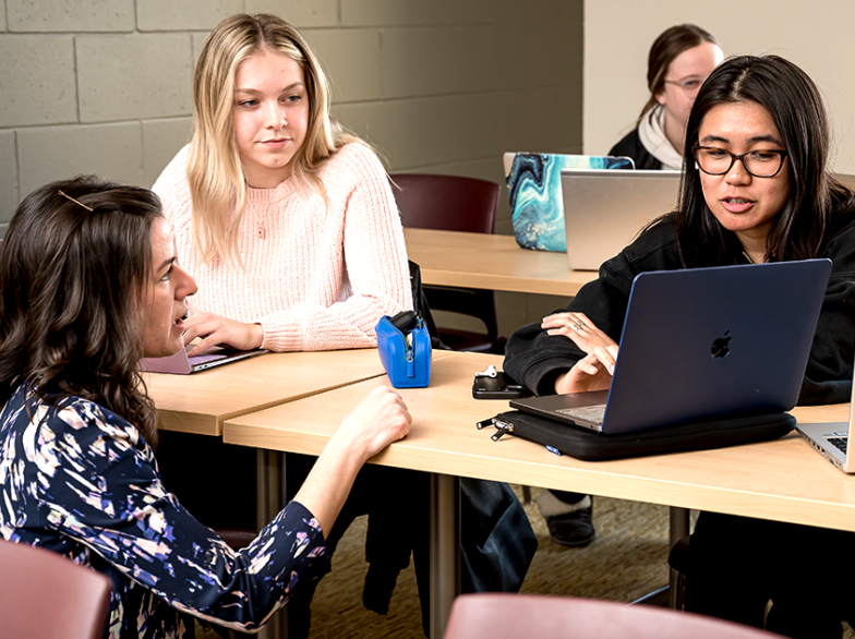 Une professeure échange avec deux étudiantes en classe à l’USB. D’autres étudiants travaillent sur leurs ordinateurs portables en arrière-plan.