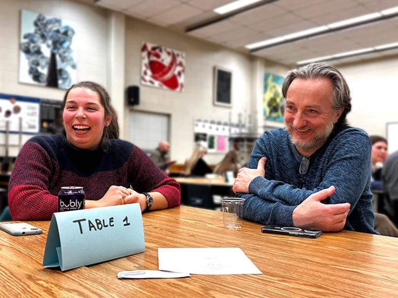 Deux personnes assises à une table dans une salle de classe sourient. Un panneau indique "Table 1". En arrière-plan, d'autres groupes sont également assis.