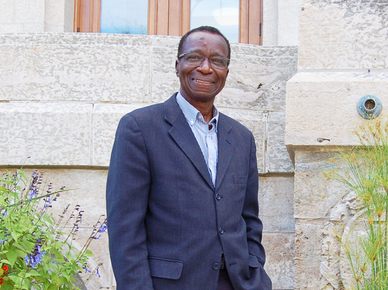 Ibrahima Diallo, souriant, en veston foncé, debout devant la façade de l’USB, entouré de plantes fleuries.