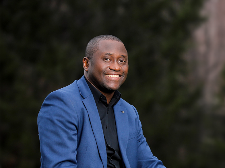 Portrait de Tadens Mpwene souriant, vêtu d’un veston bleu, assis en extérieur devant un fond flou de végétation.