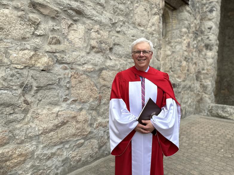 Greg Selinger, vêtu d'une toge rouge et blanche, se tient près d’un mur de pierre ancienne, un sourire sur son visage empreint de fierté.
