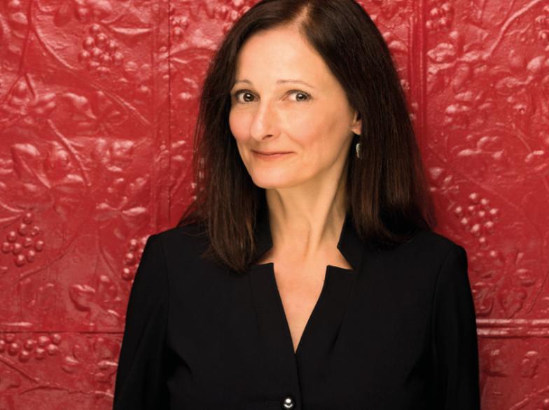 Femme souriante vêtue de noir posant devant un mur rouge texturé, regard tourné vers la caméra.
