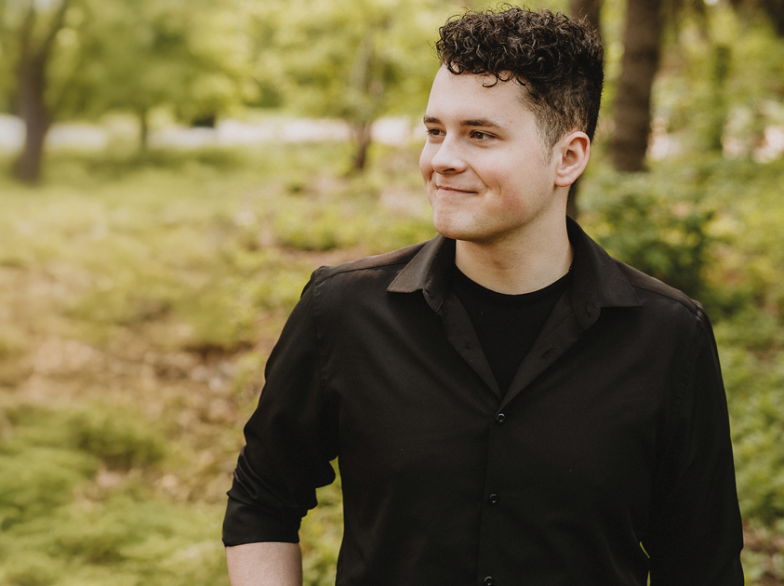 Jeune homme souriant en chemise noire debout dans un parc verdoyant, regardant vers la droite.