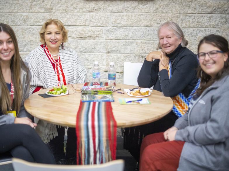 Quatre femmes assises autour d’une table ronde, dont deux Aînées métisses portant des ceintures traditionnelles, en discussion chaleureuse avec deux étudiantes.