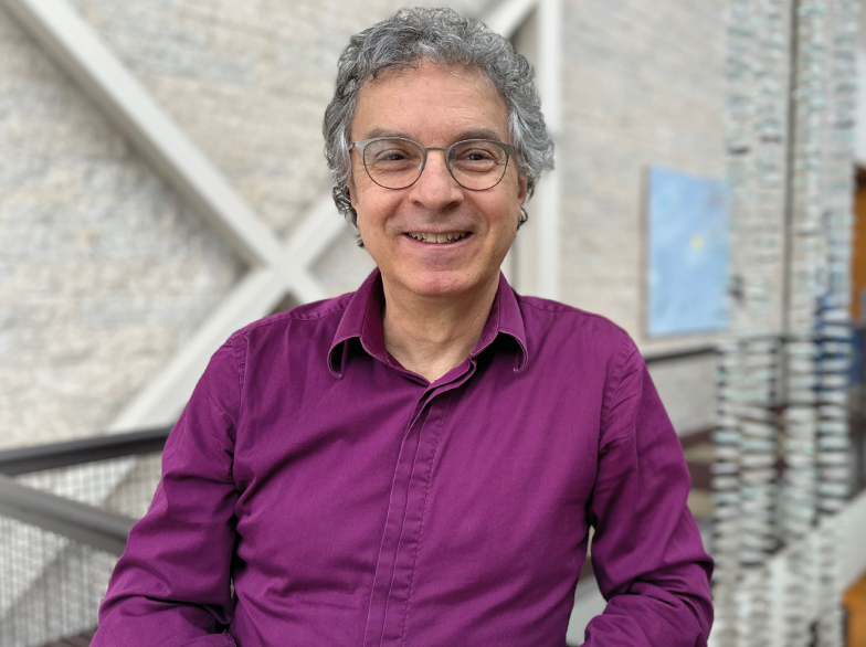 Portrait de David Alper, souriant, portant une chemise mauve, debout dans un espace intérieur lumineux de l’Université de Saint-Boniface.