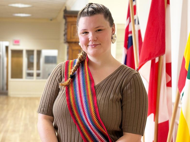 Portrait de Gabrielle Boulet, étudiante métisse portant une écharpe colorée traditionnelle, posant dans un corridor de l’Université de Saint-Boniface devant plusieurs drapeaux.