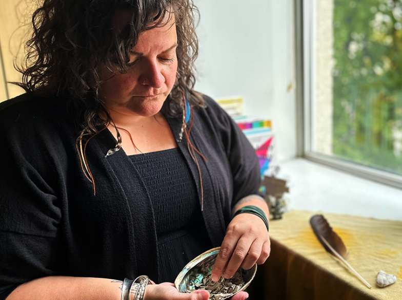 Geneviève Clément, concentrée, tient un bol d’herbes sacrées près d’une fenêtre; une plume, une plante et une robe rouge reposent sur la table.