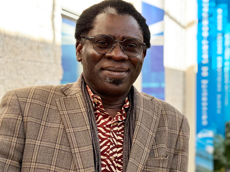 Portrait du professeur Léna Diamé Ndiaye, souriant, en veste à carreaux et chemise colorée, debout dans un corridor lumineux de l’Université de Saint-Boniface.