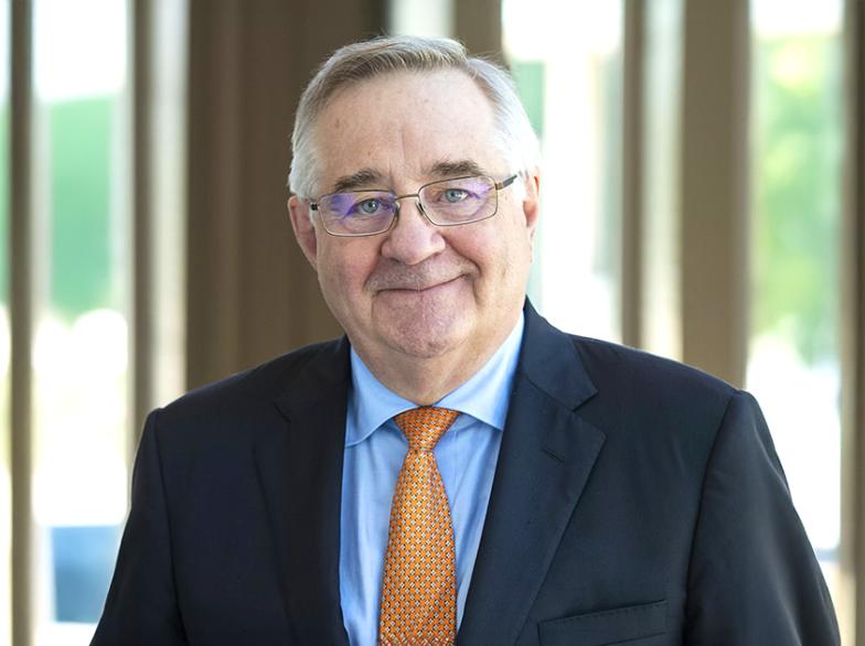 Portrait de Ronald Bisson en veston sombre et cravate orange, debout dans un corridor lumineux aux grandes fenêtres, souriant vers la caméra.