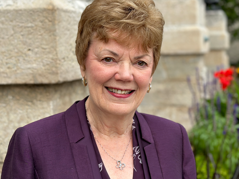 Portrait de Mariette Ferré, souriante, en veston mauve devant le campus de l’USB, avec des fleurs colorées en arrière-plan.
